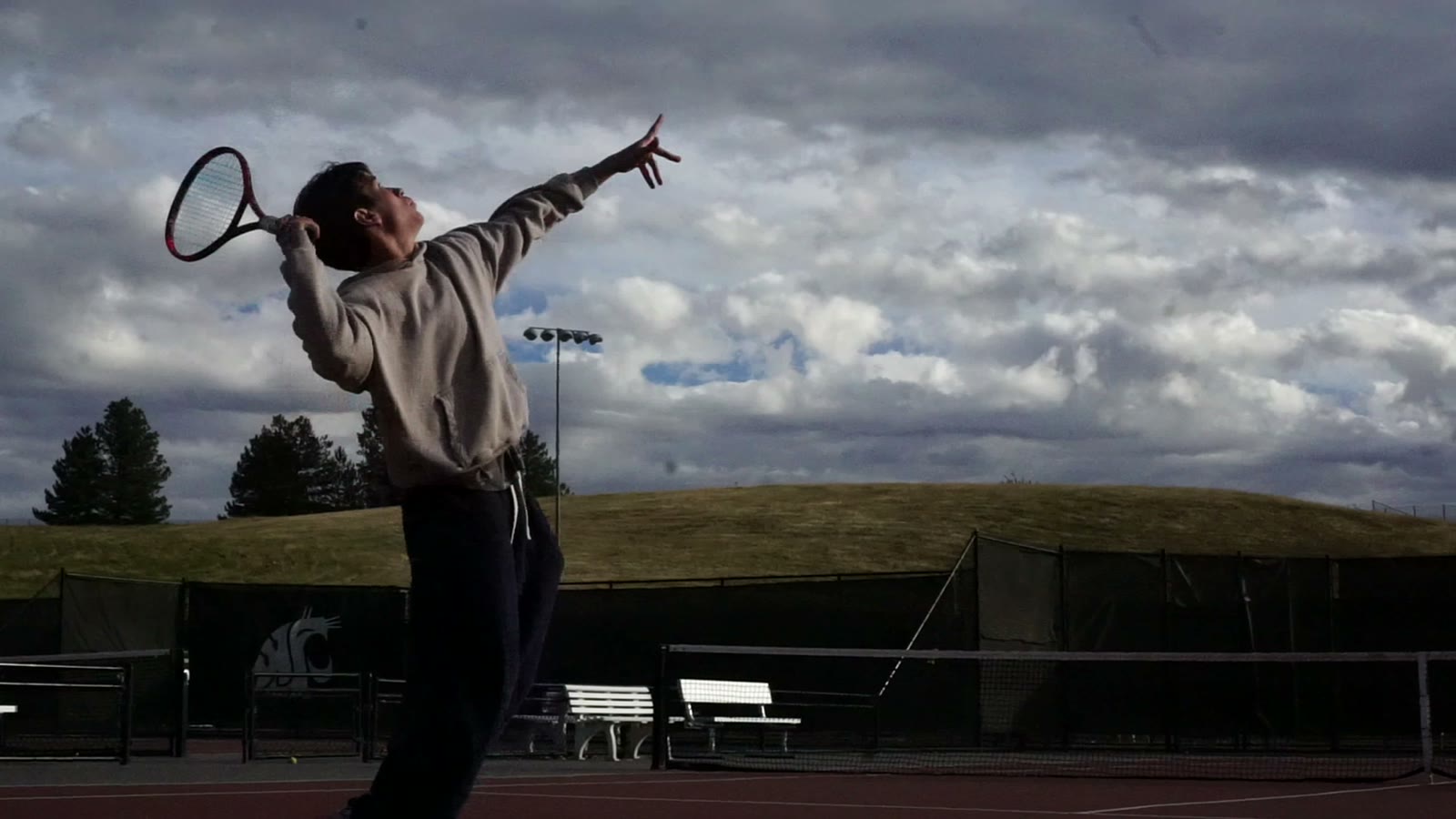 A player at full stretch on a serve under a cloudy sky